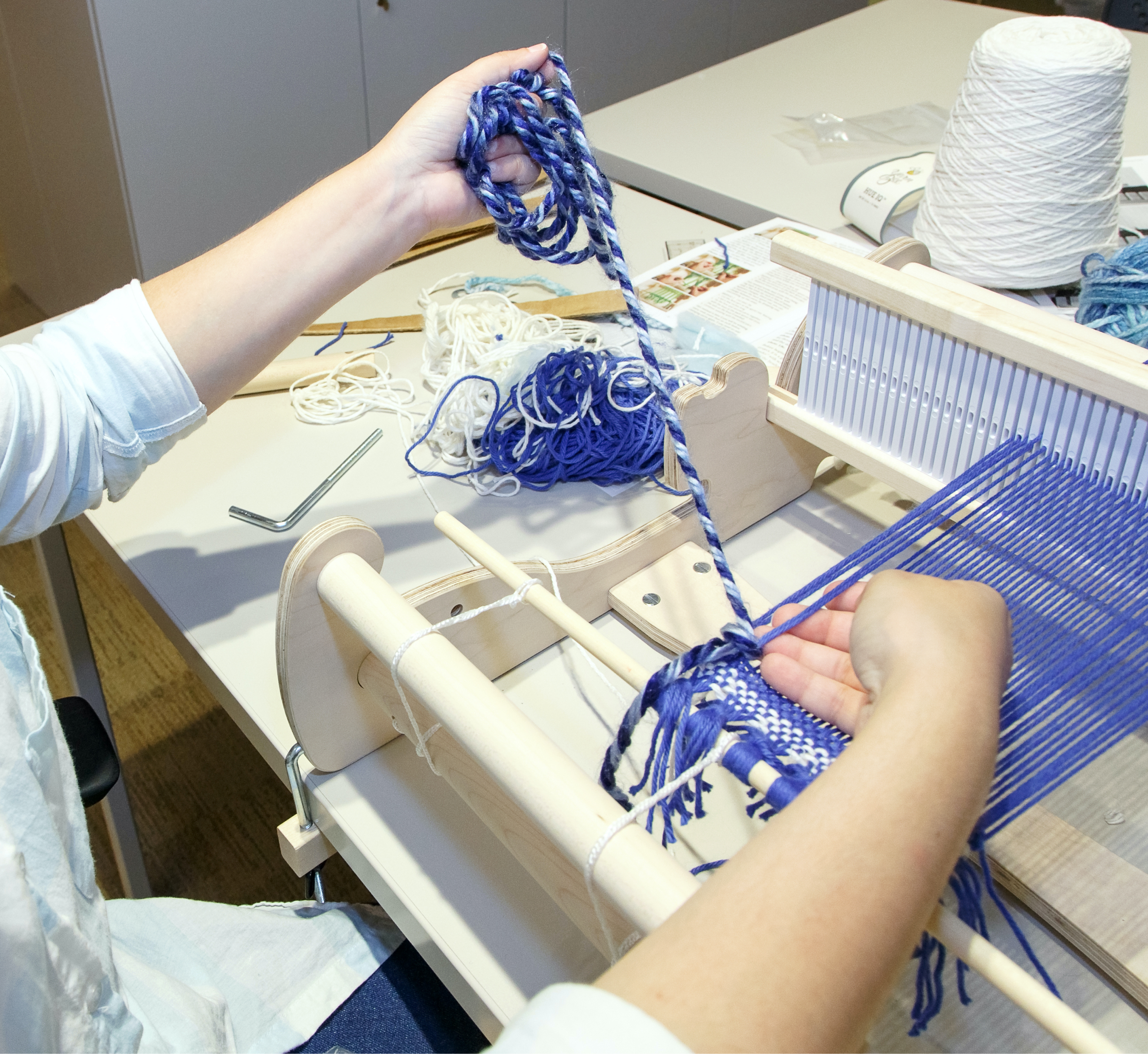 The weaver sits in front of a table top loom; one hand lifts two strand of the warp yarn, the other stretches out the yarn being looped around it.