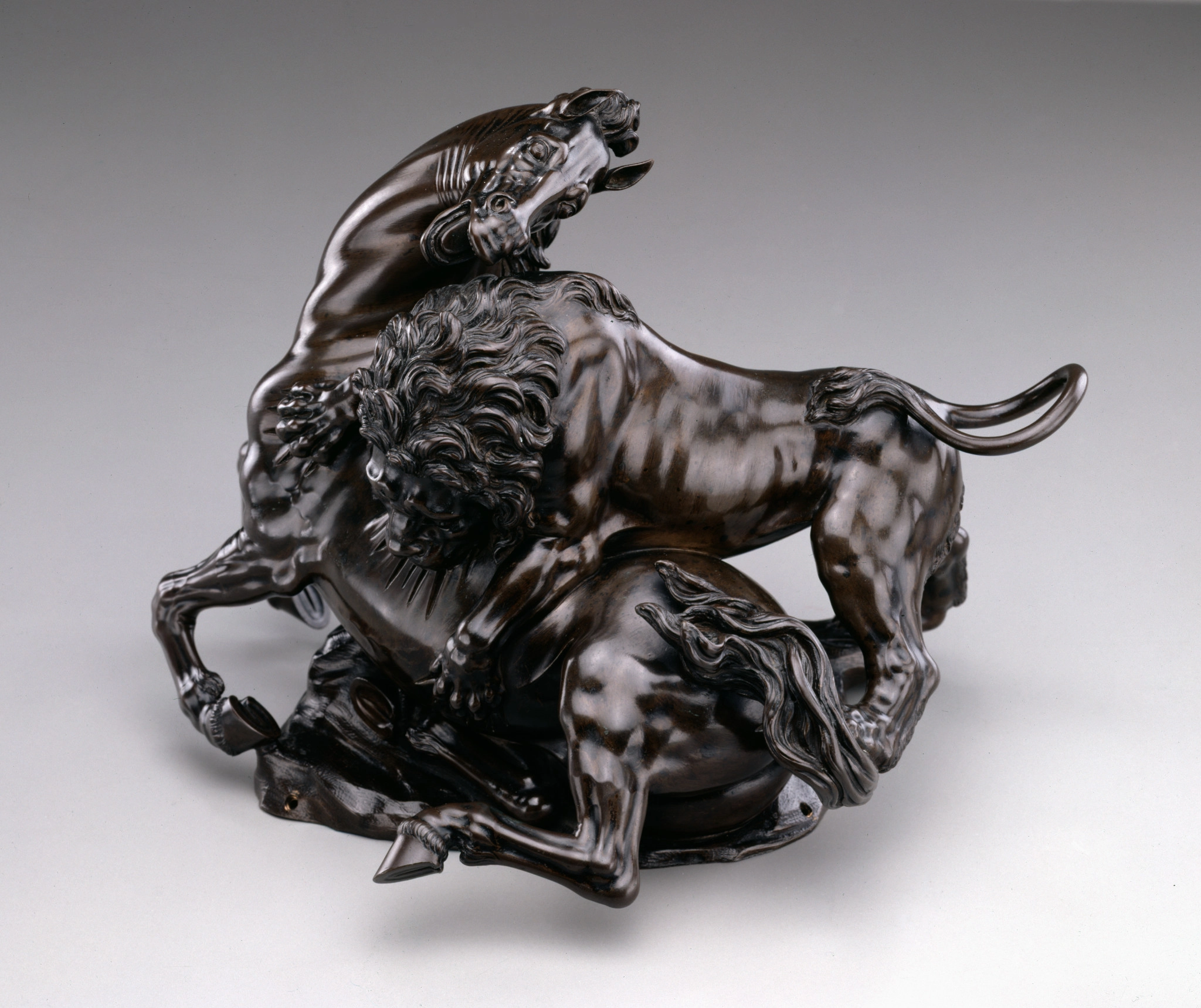 Bronze statue, deep brown color, of a lion biting into the side of a horse, who turns its head to face the lion.